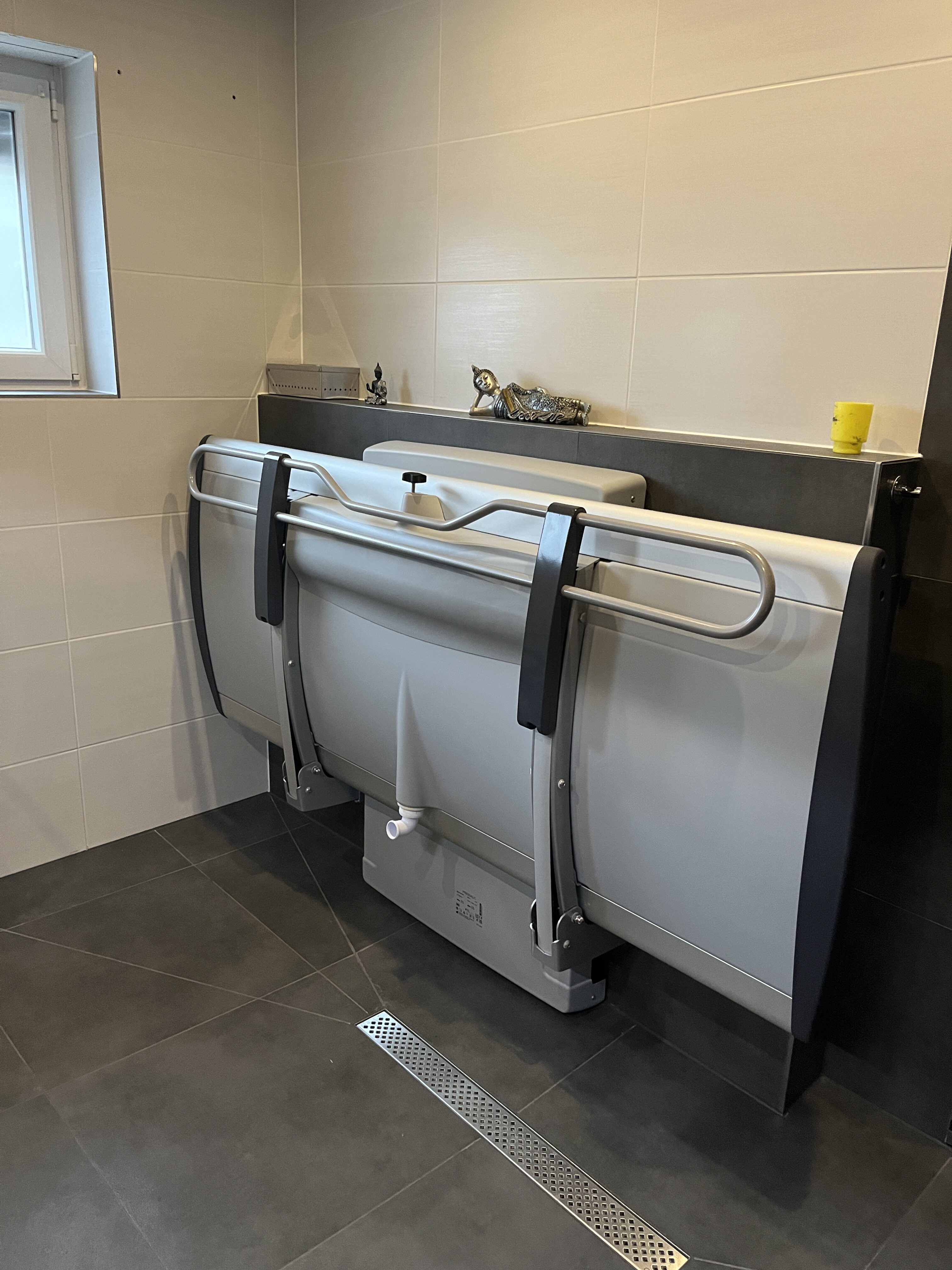 Graue Badewanne mit Klappmechanismus in modernem Badezimmer mit grauen Fliesen. Fenster im Hintergrund.